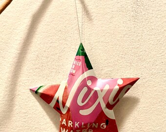May include: A pink and green star-shaped ornament made from a recycled aluminum can. The can has the text "Nixi Sparkling Water" printed on it.