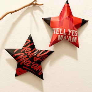 May include: Two red and black paper star ornaments with the text "Hell Yes Ma'am" on them. The stars are hanging from a white wall.