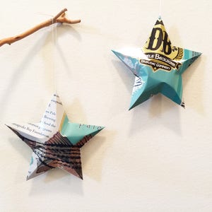 May include: Two origami stars hanging from a branch. One star is made from a book page with the text "Chesapeake Bay Foundation" and "Pale Brewing". The other star is made from a blue and gold beer can with the text "D.D.B. Pils Backbone Brewing Company".