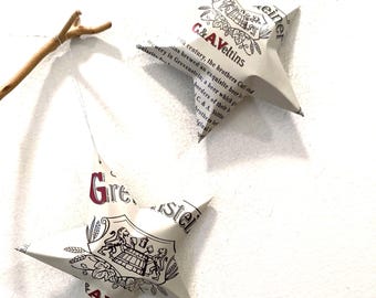 May include: Two five-pointed stars crafted from off-white paper featuring black and red printed text and a crest logo with lions. The text includes the words G & A Veltins and Grevensteiner. Each star is suspended by a thin string from a wooden branch.
