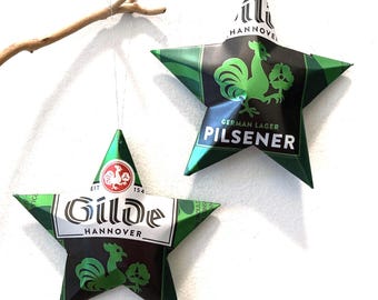 May include: Two star-shaped ornaments crafted from recycled Gilde Pilsener beer cans. The green, black, and white metal stars feature the Gilde Hannover logo, a rooster graphic, and the text German Lager Pilsener, suspended by thin silver threads.