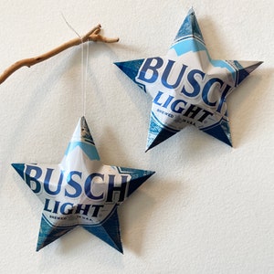 May include: Two blue and white star-shaped ornaments made from Busch Light beer cans. The stars are hanging from white string.