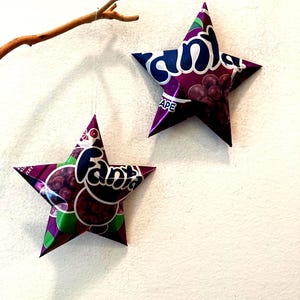 May include: Two star-shaped ornaments made from Fanta Grape soda packaging hang from a thin string. The stars are purple, green, and white, with the Fanta logo and grape imagery visible. A small branch is suspended above the stars.