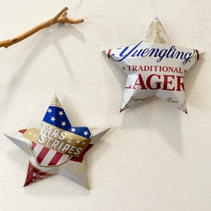 May include: Two red, white, and blue star-shaped ornaments made from aluminum beverage cans. The larger star features the text "Yuengling Traditional Lager" and the smaller star features the text "Stars & Stripes".