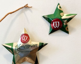 May include: Two green and gold star-shaped ornaments made from recycled beer cans. The ornaments are hanging from white string. The larger ornament has the text "Wernesgrüner Pils Legende" on it. The smaller ornament has a red "W" on it.