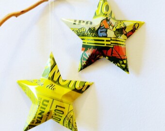 May include: Two star-shaped ornaments made from recycled metal, hanging against a white background. The stars are yellow and green, with printed text and graphics. One star hangs from a thin string, the other is positioned to the right.
