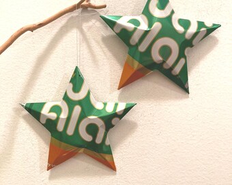 May include: Two green and orange star-shaped ornaments made from recycled aluminum cans, hanging from a branch with white string.