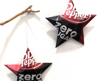 May include: Two star-shaped ornaments crafted from recycled Dr Pepper Zero Sugar aluminum cans. Each star features the red, black, and white branding with the text Dr Pepper Est. 1885 and zero sugar. One star hangs from a thin wire attached to a branch.