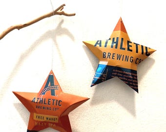 May include: Two orange star-shaped ornaments with the text "ATHLETIC BREWING CO" printed on them. One ornament also reads "FREE WAVE HAZY IPA". The stars are hanging from thin strings, with a tree branch in the background.