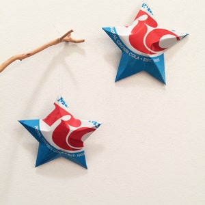 May include: Two star-shaped ornaments made from Royal Crown Cola cans hang against a white wall. The stars are blue with red and white logos. The text "Royal Crown Cola Est. 1905" is visible on the blue points of the stars. A small branch is also visible.