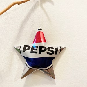 May include: A silver star-shaped ornament made from a recycled Pepsi can. The star is hanging from a white string and has the word "PEPSI" printed in black on the front.