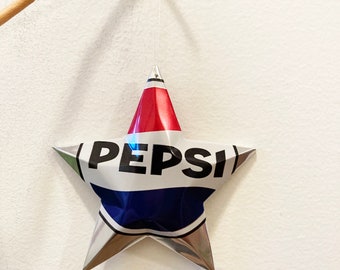 May include: A silver star-shaped ornament made from a recycled Pepsi can. The star is hanging from a white string and has the word "PEPSI" printed in black on the front.