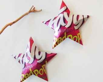 May include: Two pink and white star-shaped ornaments made from recycled Red Pop candy wrappers. The stars are hanging from a string and are against a white background.