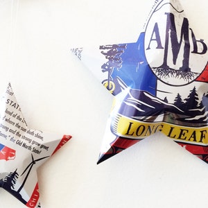 May include: Two five-pointed stars made from folded aluminum cans. The larger star is decorated with a red, white, and blue design and the text "AMD LONG LEAF". The smaller star is decorated with a white and blue design and the text "BOONE NC".