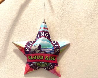 May include: A star-shaped ornament with a colorful design, hanging from a thin wire and a small branch. The ornament features the text "CLOUD RISE BREWING" in a vibrant, eye-catching font, with a landscape illustration.