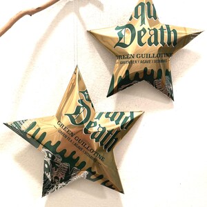 May include: Two gold-colored star-shaped ornaments made from repurposed beverage cans. The stars feature the text "Aqua Death" in green, along with "Green Guillotine" and "Green Tea x Agave x Vitamins." The ornaments are suspended by a thin string.