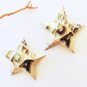May include: Two star-shaped decorations made from recycled material, hanging against a white backdrop. The stars are white with green and brown text and graphics. One star is suspended by a thin string, and the other is positioned to the right. A small branch is in the upper left corner.