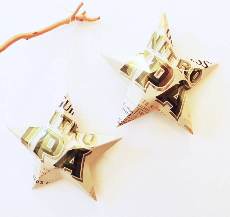 May include: Two star-shaped decorations made from recycled material, hanging against a white backdrop. The stars are white with green and brown text and graphics. One star is suspended by a thin string, and the other is positioned to the right. A small branch is in the upper left corner.