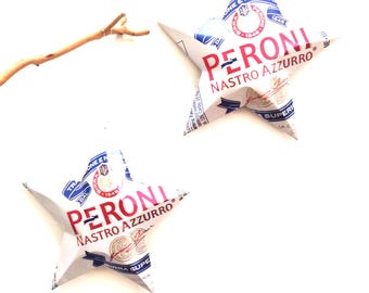 May include: Two star-shaped packages of Peroni Nastro Azzurro beer against a white background. The packages are white with red and blue text, and the brand name is prominently displayed. A small, thin, brown twig is in the upper left corner.