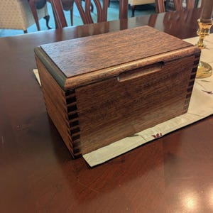 May include: A rectangular wooden box with a dark brown finish and visible finger joints at the corners. The lid features a recessed horizontal handle along the front edge, and the wood grain is prominent across the top and side surfaces.
