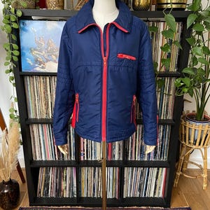 May include: A navy blue zip-up jacket featuring a bright red front zipper, a red zippered chest pocket, and two red zippered side pockets. The jacket has a fold-down collar, long sleeves with ribbed detailing at the shoulders, and a quilted texture.