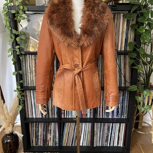 May include: A vintage brown leather jacket with a faux fur collar and a matching belt. The jacket is displayed on a mannequin, showcasing its button-up front and tailored fit. The jacket is approximately 76 cm long.