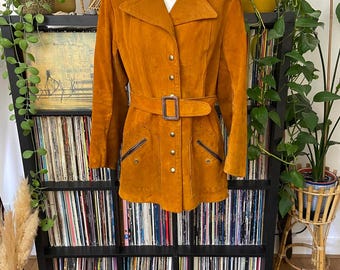 May include: A vintage mustard-colored suede jacket with a belted waist and notched collar. The jacket features a button-down front, two front pockets with zipper detailing, and a matching belt with a rectangular buckle. The jacket is displayed on a mannequin.