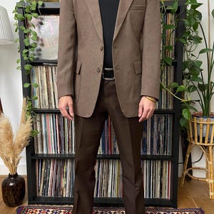 Può includere: Un blazer marrone con pantaloni marroni abbinati e una camicia nera. Il blazer ha due bottoni e revers a lancia. I pantaloni sono a gamba dritta. L'outfit è completato da scarpe in pelle nera e una cintura nera.