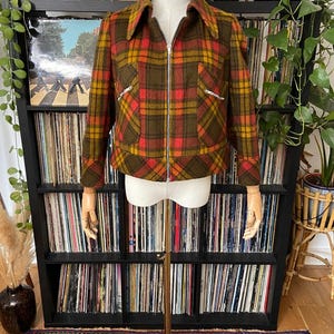 May include: A long-sleeve jacket featuring a plaid pattern in shades of olive green, mustard yellow, and bright red. The jacket has a pointed collar, a full-length silver-tone metal zipper closure, and two slanted front pockets with silver zippers.