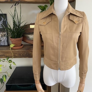 May include: A tan cropped jacket featuring a pointed collar, a full-length front zipper, and two chest pockets with button closures. The jacket has long sleeves with buttoned cuffs and a structured fit, displayed on a mannequin torso.