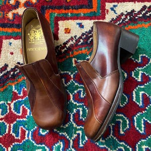 Pode incluir: Um par de sapatos de couro castanho sem atacadores com salto bloco. A palmilha interior apresenta o texto impresso a dourado Norvic Northampton England Buccaneer. Os sapatos têm um acabamento liso com costuras visíveis nos painéis laterais.