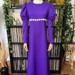May include: A long, purple dress with long sleeves and a white daisy-chain trim at the waist. The dress has a round neckline and puffed sleeves. The dress is displayed on a mannequin. The background includes plants and a shelf of vinyl records.
