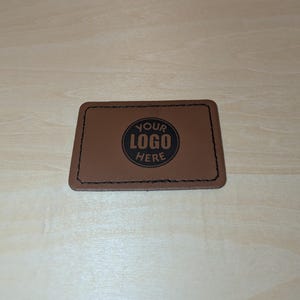 Op de afbeelding: Bruin leren patch met gestikte randen. De patch heeft een zwarte cirkel met de tekst "YOUR LOGO HERE" in wit.