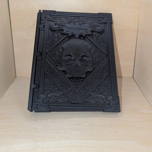 May include: A black, 3D printed book with an intricate design. The cover features a skull with a crossbones and the text "Book of Shadows".