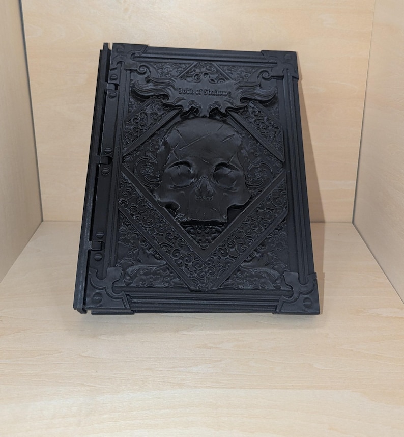May include: A black, 3D printed book with an intricate design. The cover features a skull with a crossbones and the text "Book of Shadows".