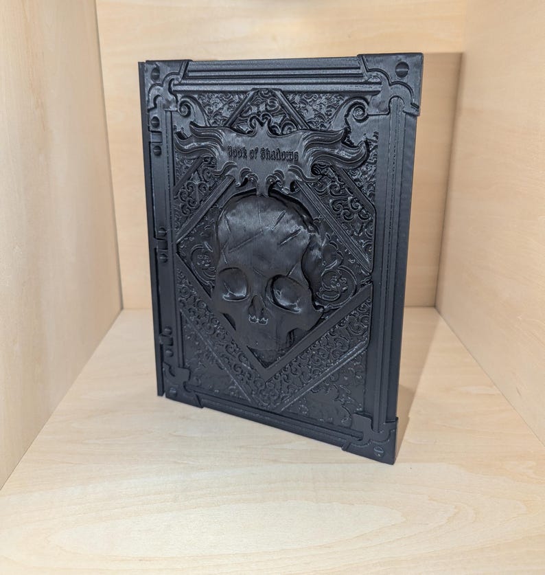 May include: A black book with an ornate, embossed cover featuring a skull and the words "Book of Shadows".
