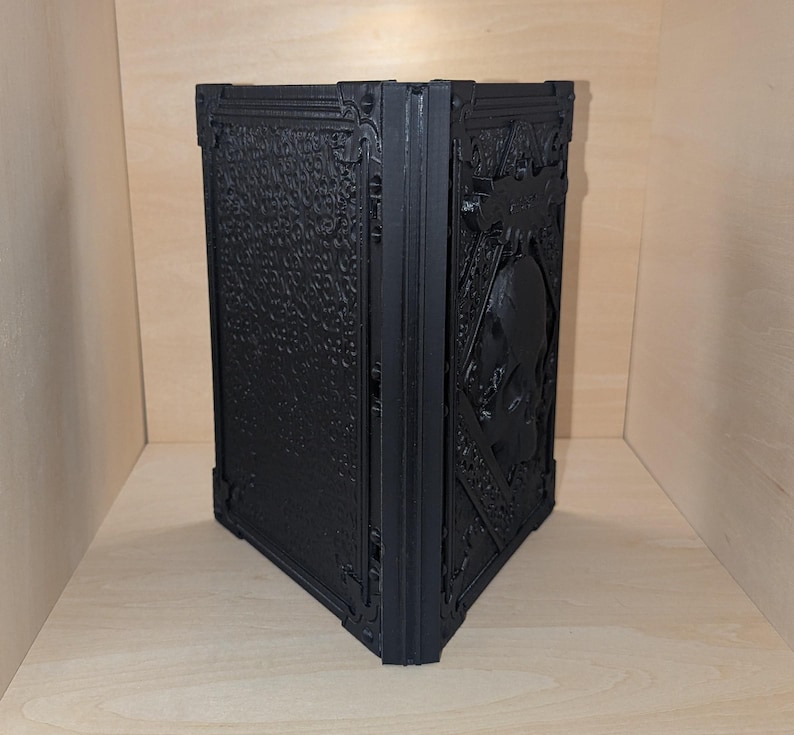 May include: A black, intricately designed, 3D printed book with a skull embossed on the front cover.