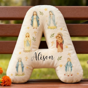 Puede incluir: Cojín en forma de letra "A" de color crema con ilustraciones de figuras religiosas y el nombre "Alison" en cursiva. El cojín presenta imágenes de la Virgen María, cruces y estrellas. El cojín está colocado sobre un banco de madera.