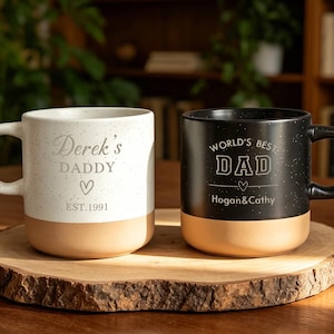 Op de afbeelding: Twee keramische mokken met gespikkelde afwerkingen en bruine bodems staan op een houten schijf. Eén mok is wit met "Derek's DADDY" en "EST. 1991" erop gedrukt. De andere is zwart met "WORLD'S BEST DAD" en "Hogan & Cathy".