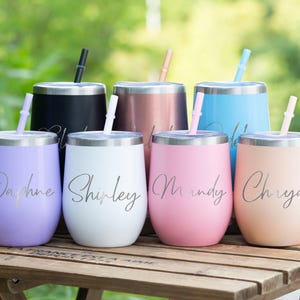 Op de afbeelding: Een verzameling geïsoleerde, steelvrije wijnglazen in diverse kleuren, waaronder zwart, roségoud, blauw, roze, lavendel, wit en perzik. Elk glas heeft een zilveren rand, een bijpassend rietje en een naam in sierlijke letters. Geplaatst op een houten tafel.