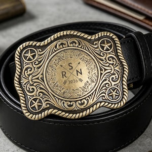 Op de afbeelding: Een zwarte leren riem met een decoratieve, antiek goudkleurige gesp. De gesp heeft een sierlijk ontwerp met sterren en een centraal cirkelvormig embleem met de initialen "S R N" en het jaar "2026".