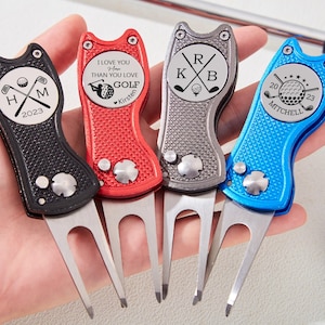 Puede incluir: Cuatro herramientas de reparación de divots de golf personalizadas en negro, rojo, plateado y azul. Cada herramienta presenta un diseño único con texto e iniciales, incluyendo una pelota de golf y palos cruzados. Las herramientas se sostienen en una mano.