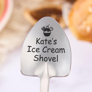 Könnte beinhalten: Ein Eiscreme-Schaufel aus Edelstahl mit einem schaufelförmigen Kopf. Der Kopf trägt den schwarzen Text "Kate's Ice Cream Shovel" und eine kleine Cupcake-Grafik. Die Schaufel ist zum Portionieren von Eiscreme gedacht.