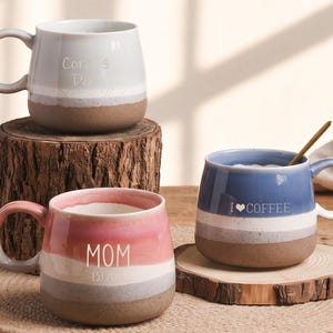 Può includere: Tre tazze in ceramica dalla forma arrotondata e smalto multicolore. Una tazza è azzurra con il testo "I LOVE COFFEE", una è rosa con "MOM EST 2023" e una è grigio chiaro con "Core's Day". Ogni tazza ha una base marrone non smaltata.