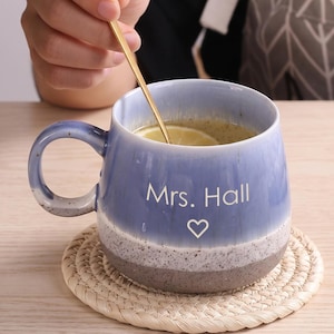 Op de afbeelding: Een keramische mok met een blauwe en grijze kleurverloopafwerking, voorzien van de tekst Mrs. Hall en een hartpictogram in het wit. De mok heeft een ronde vorm, een gebogen handvat en bevat een goudkleurige lepel in de vloeistof.