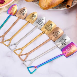 May include: A collection of shovel-shaped spoons in various metallic finishes, including gold, silver, and iridescent. Each spoon features an engraved message, such as "Don's Pastry Spoon" and "It's Coffee O'Clock". The spoons are arranged on a light-colored surface.