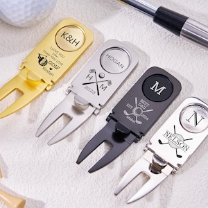 Op de afbeelding: Vier gepersonaliseerde golfdivotgereedschappen in goud, zilver en zwart. Elk gereedschap heeft een uniek ontwerp met initialen, namen en data. Eén gouden gereedschap heeft de tekst "I LOVE YOU THAN YOU LOVE GOLF Kirsten".