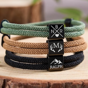 May include: Three rope bracelets in green, tan, and black, stacked on a wooden surface. Each bracelet has a black metal clasp with a unique design: initials with a heart, antlers, and a mountain scene with a name.
