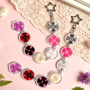 May include: Clear acrylic keychains featuring miniature record designs in various colors, connected in a chain. Each keychain has a star-shaped clasp. The records have text on them, and the background is pink.