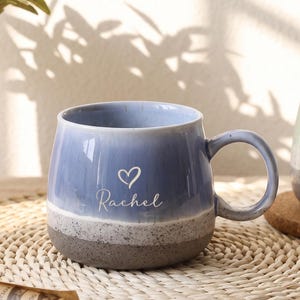 May include: A ceramic mug with a blue glazed upper section and a speckled gray base. The mug features the name Rachel in white script lettering below a small white heart icon. It has a rounded body and a curved handle on the right side.